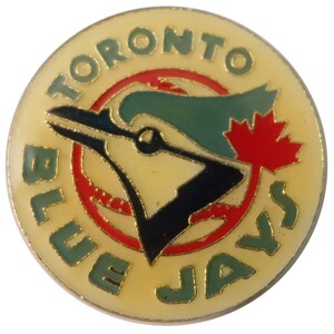 Può includere: Uno spillo dorato con un logo di un ghiandaia azzurra. Il logo presenta un ghiandaia azzurra con una foglia d'acero rossa sulla testa. Le parole "Toronto Blue Jays" sono scritte intorno al logo.