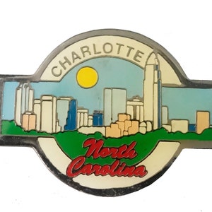 May include: A silver metal pin with a colorful image of the Charlotte, North Carolina skyline. The pin features a blue sky with a yellow sun, a green grassy area, and the text "Charlotte" and "North Carolina".