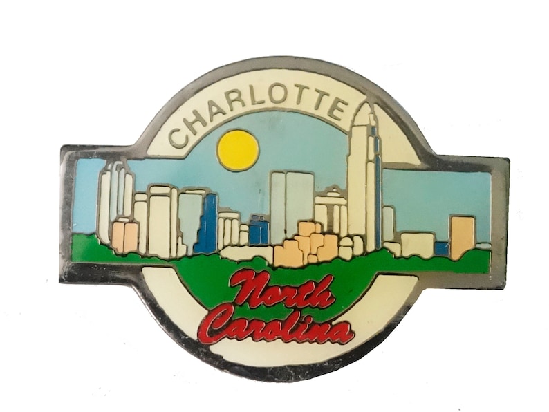 May include: A silver metal pin with a colorful image of the Charlotte, North Carolina skyline. The pin features a blue sky with a yellow sun, a green grassy area, and the text "Charlotte" and "North Carolina".