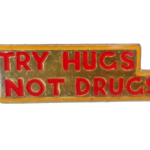 May include: A gold-colored metal pin with a yellow border and red lettering that reads "TRY HUGS NOT DRUGS".