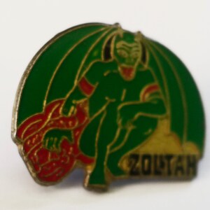 May include: A green and gold enamel pin featuring a cartoon devil with red horns and a red tail. The devil is sitting on a gold mound with the word "ZOLTAN" written on it.