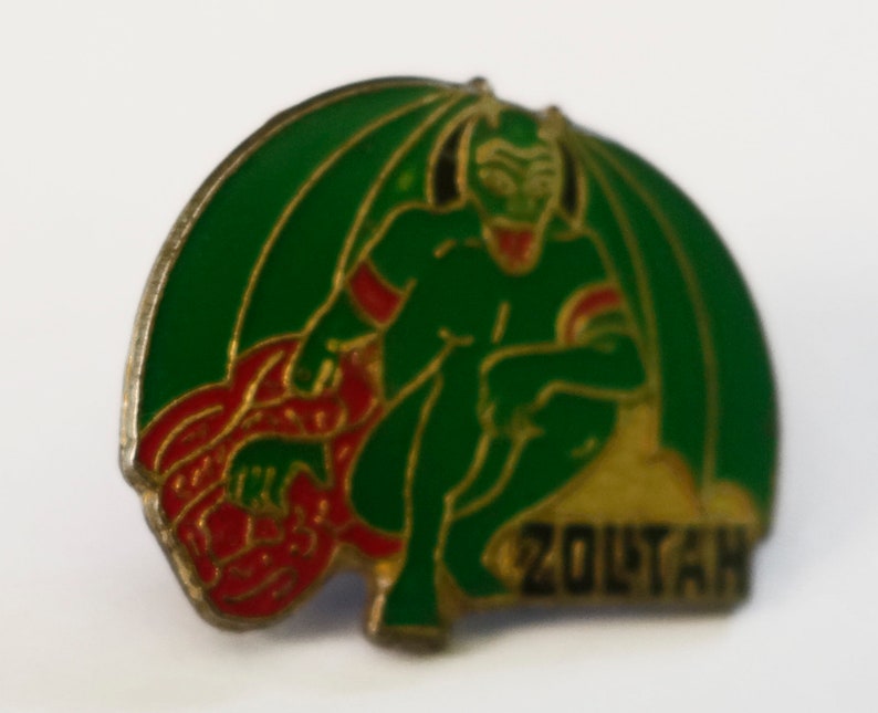 May include: A green and gold enamel pin featuring a cartoon devil with red horns and a red tail. The devil is sitting on a gold mound with the word "ZOLTAN" written on it.