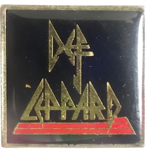 May include: A gold metal pin with a black background featuring the band name "Def Leppard" in gold lettering. The band's logo is a stylized lightning bolt with a red stripe at the bottom.