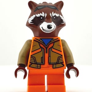 Puede incluir: Una minifigura de Lego Rocket Raccoon. La figura tiene una cabeza de mapache marrón con marcas blancas y gafas. Viste un mono naranja con una chaqueta marrón y brazos marrones. Las manos son de color marrón oscuro.