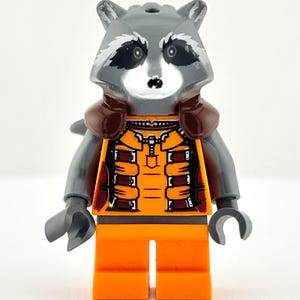 Puede incluir: Una minifigura de Lego de Rocket Raccoon de la serie Guardianes de la Galaxia. La figura tiene una cabeza de mapache gris con marcas negras, vistiendo un mono naranja con detalles marrones. La figura está sobre una superficie blanca.