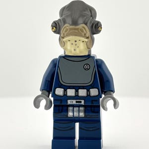 May include: A Lego minifigure of a Quarren character, featuring a dark blue uniform with gray accents. The figure has a gray head with distinctive tentacles and a tan face. The figure is standing against a white background.