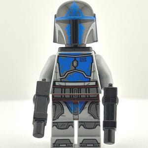 May include: A Lego minifigure of a Mandalorian warrior. The figure has a gray and blue color scheme, with a helmet and armor. It is holding two black blasters. The figure is standing against a white background.