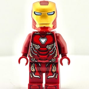 Puede incluir: Una figura de Lego de Iron Man roja y dorada. La figura tiene un cuerpo rojo con detalles dorados y un casco dorado con ojos azules. La figura está de pie sobre un fondo blanco.