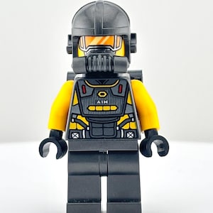 May include: A Lego minifigure of a character in a black helmet, mask, and vest with yellow accents. The vest has a design with the word "AIM" on it. The figure has yellow arms and black hands and legs.