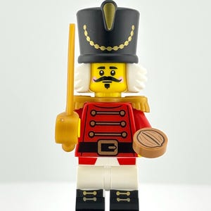 以下が含まれることがあります： 赤色のジャケット、白いズボン、黒いブーツを履いた、レゴのくるみ割り人形のフィギュア。 金色の装飾が施された黒い帽子、黄色の剣、茶色のオブジェクトを持っています。 黒い台座の上に立っています。 高さ約4cm。