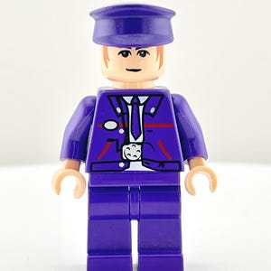 Puede incluir: Una figura de Lego morada de un conductor con sombrero y uniforme a juego. La figura tiene una corbata blanca, una insignia y detalles rojos en la chaqueta. La figura está sobre una superficie blanca.