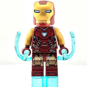 Puede incluir: Una figura de Lego Iron Man con un casco dorado y una armadura roja. La figura tiene efectos de energía azules alrededor de las manos y los pies. La figura está sobre una superficie blanca.