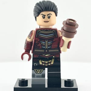 May include: A Lego minifigure with black hair and a stern expression. The figure wears a red and gold patterned outfit and holds a brown accessory. It stands on a black base against a white background. The figure is a collectible toy.