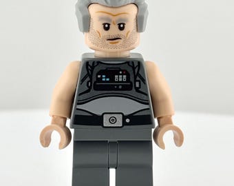 May include: A Lego minifigure of an older man with gray hair and a matching beard. He wears a gray outfit with a black design on the chest and a silver belt buckle. The minifigure has a neutral expression and is standing upright.