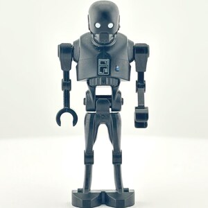 May include: A black K-2SO action figure from the Star Wars franchise. The figure has a rounded head with two white eyes, a rectangular torso, and articulated limbs. The figure is standing on a white surface.
