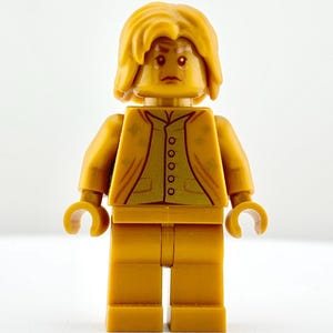 May include: A gold-colored Lego minifigure with a serious expression. The figure has a yellow hairstyle and a button-down shirt with a detailed design. The minifigure is standing upright with its arms at its sides.