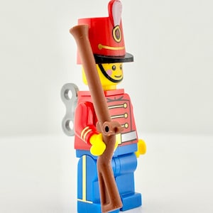 以下が含まれることがあります： プラスチック製の兵隊のおもちゃのフィギュア。兵士は赤いジャケット、青いズボン、高い赤い帽子を着用しています。茶色のライフルを持ち、背中には巻き上げキーが付いています。兵士は笑顔の黄色い顔をしています。