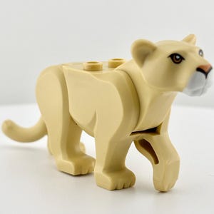 May include: A tan-colored plastic lioness toy figure. The toy has a detailed head with brown eyes, a pink nose, and a white muzzle. The lioness is in a walking pose, with its tail extended.