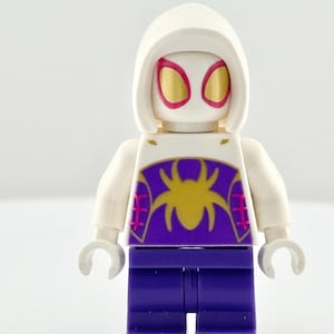 Op de afbeelding: Een Lego Spider-Gwen minifiguur. De figuur heeft een witte kap met gouden en roze oogdetails. De torso is wit met een paars spinnenembleem en roze accenten. De broek is paars en de armen wit.