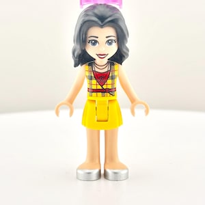 Könnte beinhalten: Eine Lego Friends Mini-Puppe mit schwarzen Haaren, blauen Augen und einem rosa Heiligenschein. Die Figur trägt ein gelbes kariertes Oberteil und einen Rock mit silbernen Schuhen. Die Figur steht aufrecht.
