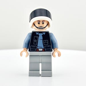 May include: A Lego minifigure of a man wearing a white helmet with a black stripe, a black vest, a blue shirt, and gray pants. The minifigure has a neutral expression and is standing upright.