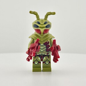 Op de afbeelding: Een plastic minifiguur in de vorm van een insectachtig wezen met olijfgroene huid, donkerrode accenten op de romp en benen, en grote zwarte ogen. Het figuurtje heeft twee antennes op het hoofd en houdt twee rode, klauwachtige accessoires in de handen.