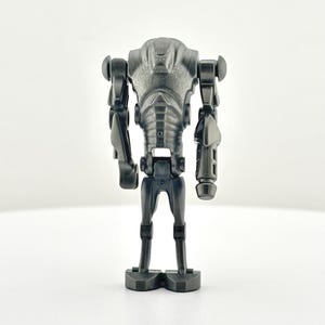 May include: A gray battle droid toy figure with a detailed, mechanical design. The figure has a rounded head, articulated arms, and a segmented torso. It stands on two feet and is set against a white background.
