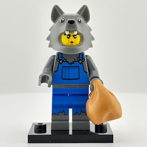 May include: A Lego minifigure of a wolf in a gray wolf head costume, blue overalls, and a tan sack. The minifigure is standing on a black base. The wolf head has a yellow face.
