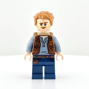 May include: A Lego minifigure of a man with light brown hair and a short beard. He wears a brown vest over a blue shirt, blue jeans, and a brown backpack. The figure has a neutral expression and is standing against a white background.