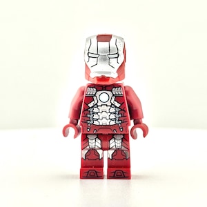 Puede incluir: Una minifigura de Lego de Iron Man roja y plateada. La figura tiene un cuerpo rojo con detalles de armadura plateada en el pecho, las piernas y los brazos. La cabeza es un casco plateado con una placa frontal roja. La figura está de pie.