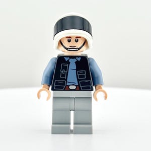 May include: A Lego minifigure of a character wearing a black and white helmet, a blue shirt, a black vest, and gray pants. The minifigure has a neutral expression and is standing against a white background.