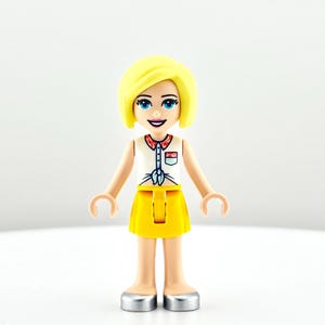 May include: A Lego Friends mini-doll figure with short yellow hair, blue eyes, and a smiling expression. She wears a white top with a red collar and a yellow skirt. Her shoes are silver. The figure is standing against a white background.