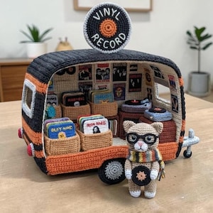 May include: A crocheted miniature camper van featuring a black and orange exterior with a circular sign on top reading VINYL RECORD. The interior displays tiny record crates and players. A small crocheted cat figure wearing glasses and a scarf stands in front.