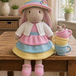 Puede incluir: Una muñeca de conejo tejida con un sombrero de rayas pastel con una flor, un cárdigan blanco y un vestido de capas en rosa, azul y amarillo. El conejo lleva zapatos rosas y está sentado junto a una pila de tres pequeñas tazas tejidas con un ratón.
