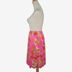 K&ouml;nnte beinhalten: Knielanger Rock mit einem lebendigen, abstrakten Blumenmuster in Pink-, Orange- und Gr&uuml;nt&ouml;nen. Der Rock wird auf einer Schaufensterpuppe pr&auml;sentiert und hat einen gewellten Saum und einen Schlitz vorne.