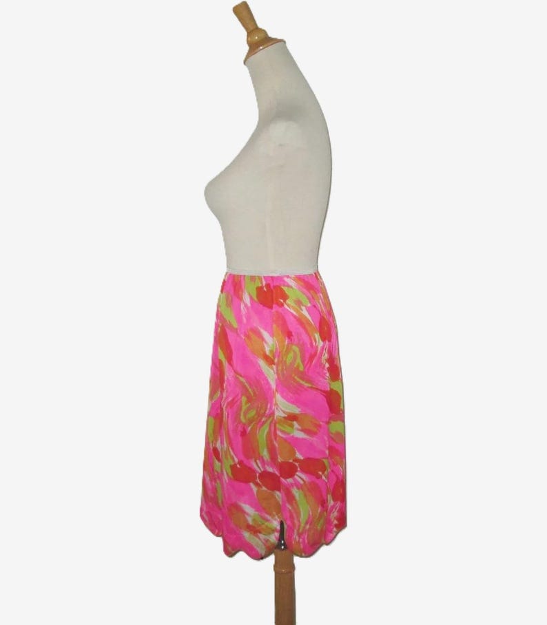 K&ouml;nnte beinhalten: Knielanger Rock mit einem lebendigen, abstrakten Blumenmuster in Pink-, Orange- und Gr&uuml;nt&ouml;nen. Der Rock wird auf einer Schaufensterpuppe pr&auml;sentiert und hat einen gewellten Saum und einen Schlitz vorne.