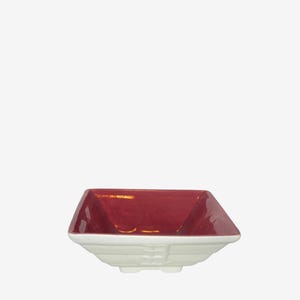 May include: A square ceramic bowl featuring a deep red interior and a contrasting white exterior. The bowl has a stepped, tiered base design with four small feet, providing a stable foundation for this functional kitchen serving piece.