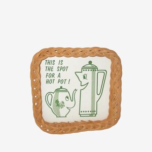 May include: A square, woven trivet with a white center featuring green cartoon teapots and the text "THIS IS THE SPOT FOR A HOT POT!". The trivet has a light brown, woven border.