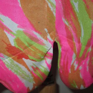 K&ouml;nnte beinhalten: Nahaufnahme eines Stoffes mit lebendigem, abstraktem Muster. Das Design zeigt kr&auml;ftige Striche in Neonrosa, Orange, Gr&uuml;n und Wei&szlig;. Der Stoff ist wahrscheinlich ein Kleidungsst&uuml;ck, mit einem Schlitz am Saum.