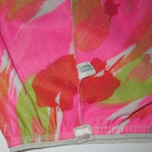 K&ouml;nnte beinhalten: Nahaufnahme eines Kleidungsst&uuml;cks mit leuchtendem Blumenmuster in Pink, Orange, Gr&uuml;n und Wei&szlig;. Der Stoff hat eine strukturierte Optik. Ein kleines Etikett tr&auml;gt die Aufschrift "LORRA PERFECT FIT". Der elastische Rand des Kleidungsst&uuml;cks ist sichtbar.