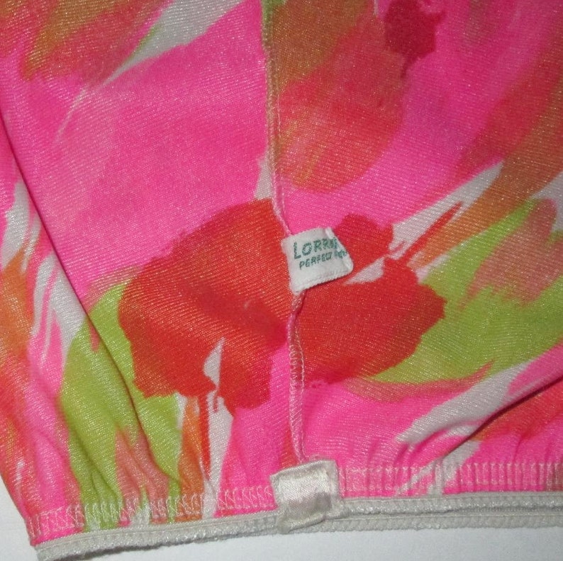 K&ouml;nnte beinhalten: Nahaufnahme eines Kleidungsst&uuml;cks mit leuchtendem Blumenmuster in Pink, Orange, Gr&uuml;n und Wei&szlig;. Der Stoff hat eine strukturierte Optik. Ein kleines Etikett tr&auml;gt die Aufschrift "LORRA PERFECT FIT". Der elastische Rand des Kleidungsst&uuml;cks ist sichtbar.