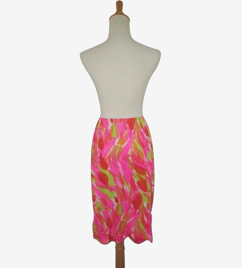 K&ouml;nnte beinhalten: Ein lebendiger, knielanger Rock mit einem farbenfrohen, abstrakten Muster. Der Rock hat eine hohe Taille und einen gewellten Saum. Das Design umfasst rosa, gr&uuml;ne und orangefarbene Striche auf wei&szlig;em Hintergrund. Der Rock wird auf einer Schaufensterpuppe pr&auml;sentiert.