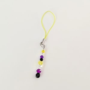 May include: A beaded charm with a yellow cord and silver-colored metal clasp. The charm features a string of colorful beads in yellow, purple, black, and clear. The beads are faceted and have a shiny appearance.