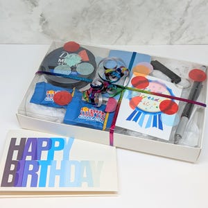 Puede incluir: Un set de regalo de cumpleaños en una caja transparente, que incluye una tarjeta que dice "HAPPY BIRTHDAY" en letras degradadas azules y moradas. La caja contiene un producto Dove Men+Care, barras de chocolate azules, un cuaderno, un bolígrafo y una tarjeta de cumpleaños.