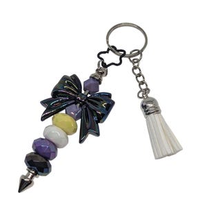 May include: A keychain featuring a dark iridescent bow, a beaded strand with purple, yellow, and white beads, and a white tassel. The keychain has a silver-tone metal ring and accents, and a pointed end.
