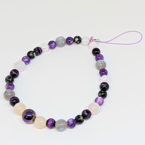 May include: A purple, white, and black beaded phone charm with a lavender lanyard. The beads are round and have a variety of patterns.
