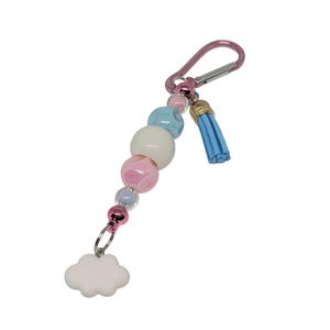 May include: A beaded keychain with a pink carabiner clip. The keychain features a strand of pastel-colored beads, including pink, blue, and white, and a small blue tassel. A white cloud charm hangs at the bottom.
