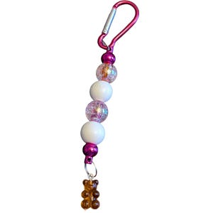 May include: A keychain featuring a pink carabiner attached to a series of alternating white spherical beads and clear crackle-textured beads with pink accents. At the bottom, a small, translucent brown bear-shaped charm hangs from a silver ring.