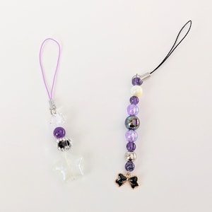 May include: Two beaded phone charms. One has a purple loop, clear star, and purple and silver beads. The other has a black cord, purple, iridescent, and silver beads, and a black bow charm.