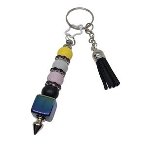 Könnte beinhalten: Ein Schlüsselanhänger mit einem silbernen Ring und einem dekorativen Perlenanhänger. Der Anhänger hat einen schillernden Würfel, schwarze, rosa, graue und gelbe Perlen und eine schwarze Quaste. Die Perlen sind durch silberne Akzente getrennt.